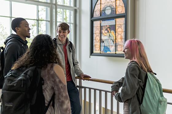 Photo of a group of four Chatham University students laughing after class in brightly lit academic building with a stained glass window. 