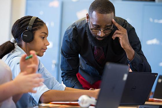 Photo of a professor leaning over a student working on a laptop and wearing black headphones in a computer lab. 